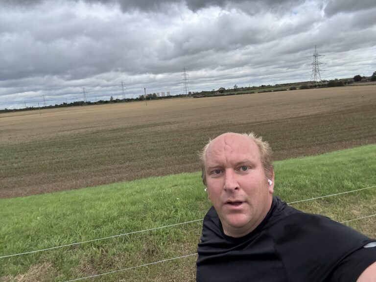 Chris taking a selfie beside open fields during a run, beneath a cloudy sky.